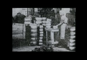 Czarno-białe zdjęcie odlewni w Grudziądzu. W środku stosy garnków, po lewej pracownica odlewni