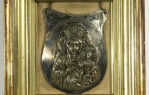 Fotografia srebrnego ryngrafu z wizerunkiem Matki Boskiej z Dzieciątkiem, oprawiony w ramę barwy złotej.