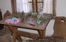 Kolorowa fotografia – drewniany stół, na nim gałązki bazi, gotowe palmy, nóż. W tle okno, obok drewniane krzesło