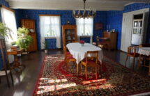 Kolorowa fotografia pokoju – na środku stół, pod ścianami inne meble z jasnobrązowego drewna, na podłodze dywan. Na ścianach niebieska tapeta