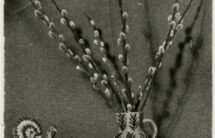 Fotografia czarno-białej pocztówki – półka przykryta wyszywaną serwetą, na niej wazon z baziami, pisanki i gliniany kogut.