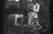 Czarno-biała fotografia: młoda kobieta z niemowlęciem na ręku, obok mały chłopiec i wózek dziecięcy. Scena w ogrodzie, w tle wiejskie chaty