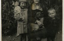 Czarno-biała fotografia: kobieta siedzi na krześle z psem na kolanach, obok stoi dziewczynka i chłopiec.
