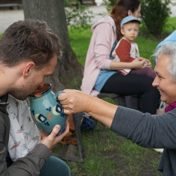 Kobieta podkłada chłopcu siedzącemu na kolanach mężyczny gliniany dzbanek. W tle kobieta z dzieckiem na kolanach.