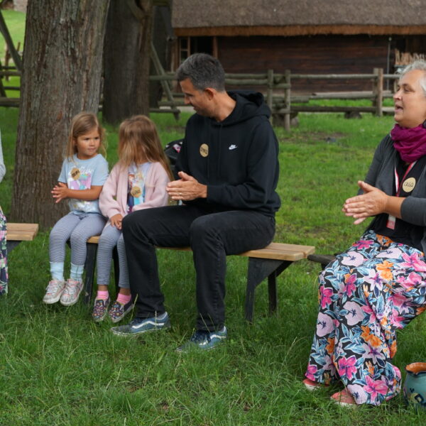 Kobieta oraz rodzina: kobieta, meżczyzna i dwie dziewczynki siedzą na drewnianych ławkach i klaszczą w dłonie 