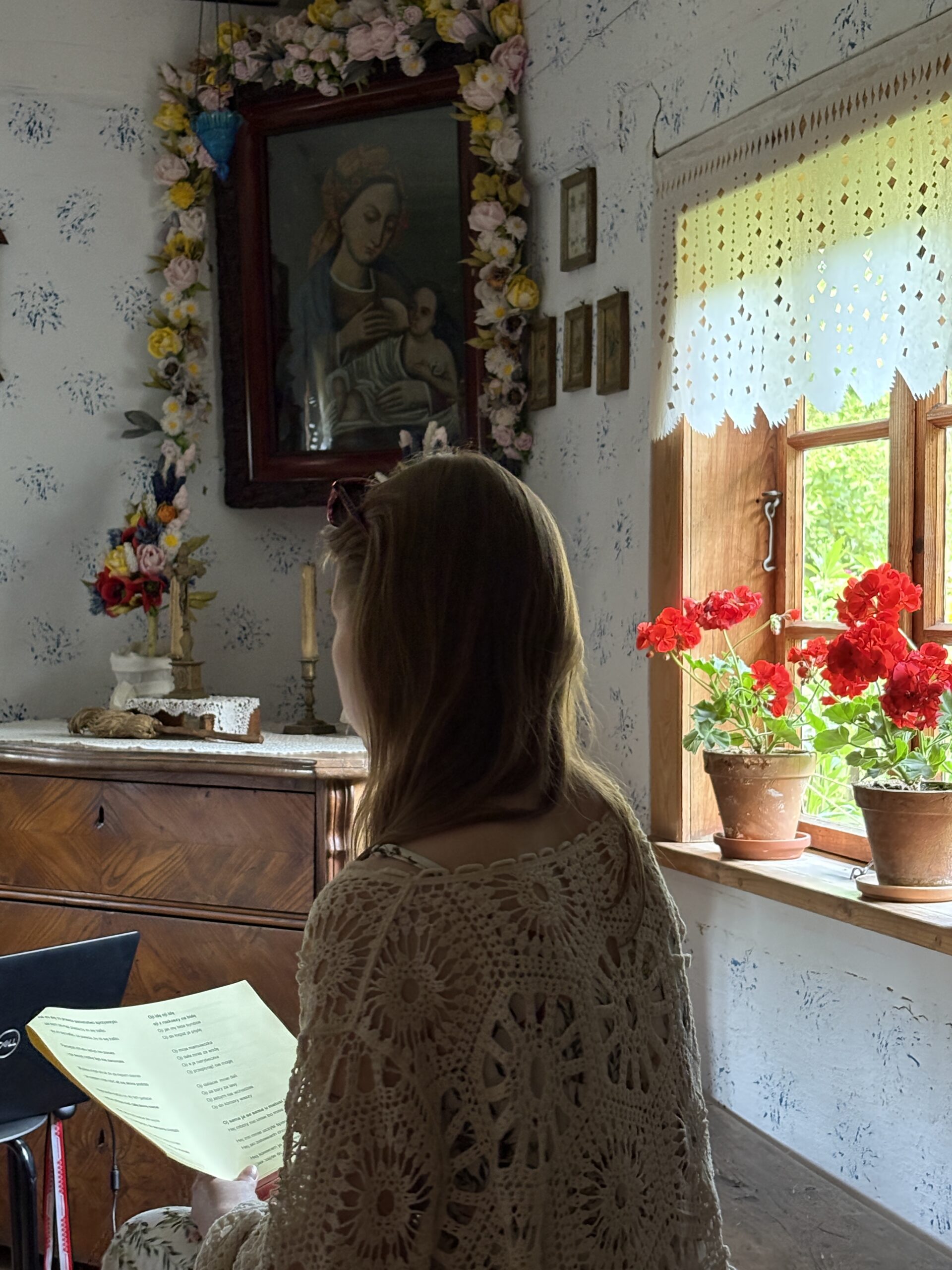 Kobieta siedzi tyłem w starej chałupie kujawskiej, tzyma kartkę ze słowami pieśni