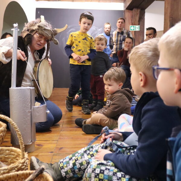 Rodziny z małymi dziećmi zebrane w sali muzeum. Wśród nich kobieta w czapce z rogami kozy. 
