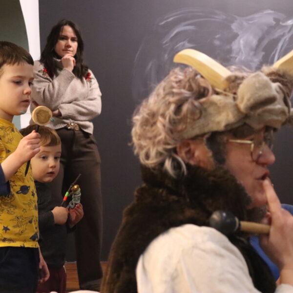 Rodziny z małymi dziećmi zebrane w sali muzeum. Wśród nich kobieta w czapce z rogami kozy. 