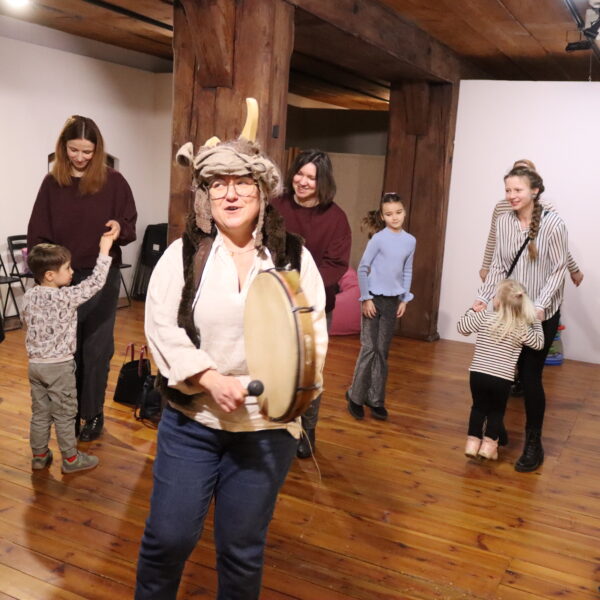 Rodziny z małymi dziećmi zebrane w sali muzeum. Wśród nich kobieta w czapce z rogami kozy. 