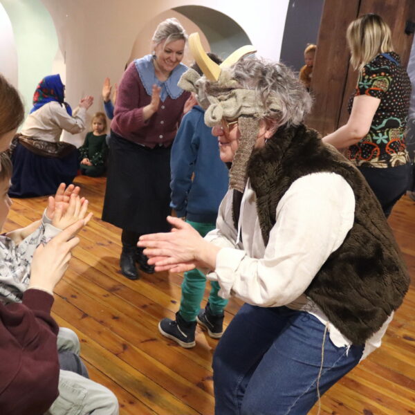 Rodziny z małymi dziećmi zebrane w sali muzeum. Wśród nich kobieta w czapce z rogami kozy. 