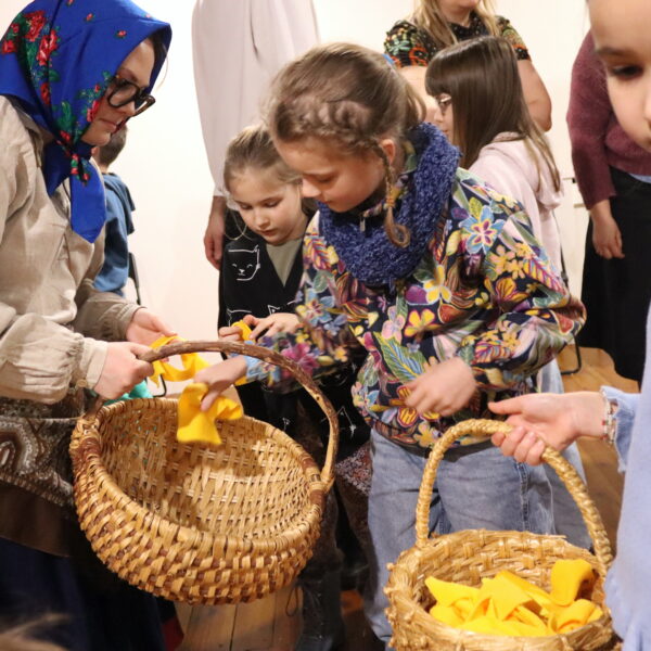 Kobieta w niebieskiej, haftowanej chuście na głowie podaje dzieczynce wiklinowy koszyk 