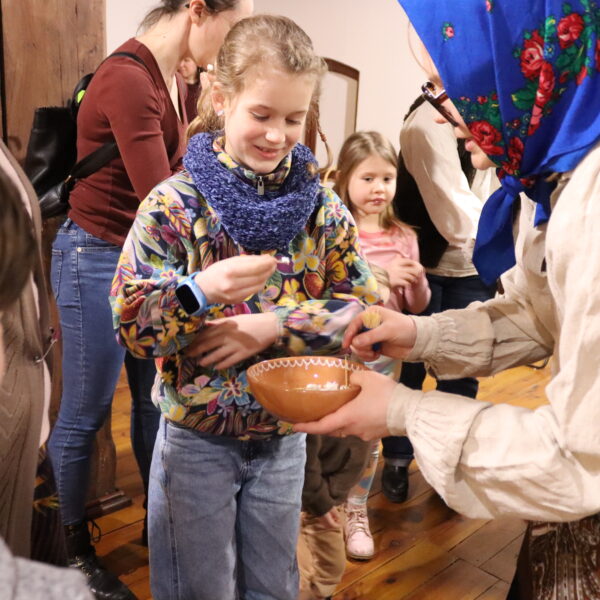 Kobieta w niebieskiej, haftowanej chuście na głowie podaje dzieczynce miskę. 