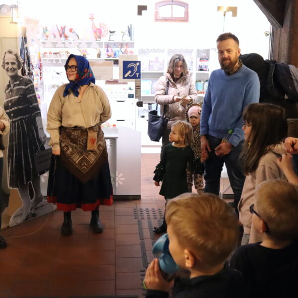 Kobieta z niebieską, haftowaną chustą na głowie stoi przed dziećmi i dorosłymi. 