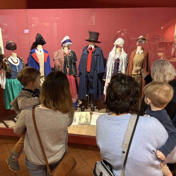 Dwie kobiety stoją przed witryną z tradycyjnymi strojami kujawskimi. Na rękach każda z nich trzma dziecko. 