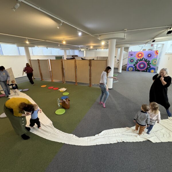 Kobiety z dziećmi bawią się w sali na zielonej wykładzinie, na podłodze rozłożony jest długi kawałek materiału, po którym biegają. 