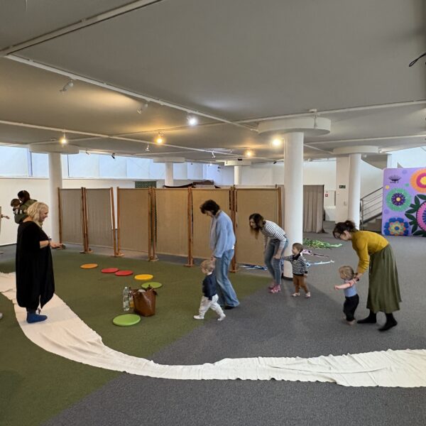 Kobiety z dziećmi bawią się w sali na zielonej wykładzinie, na podłodze rozłożony jest długi kawałek materiału, po którym biegają. 