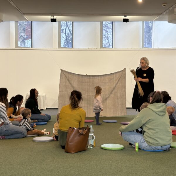 Kobiety z dziećmi siedzą na zielonej wykładzinie, obserwując kobietę z bukietem lnu stojącej przy zawieszonej lnianej tkaninie 