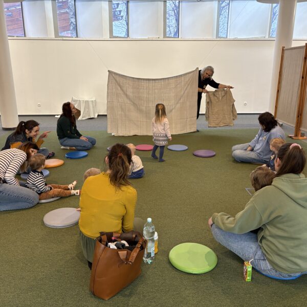 Kobiety z dziećmi siedzą na zielonej wykładzinie, obserwując kobietę z bukietem lnu stojącej przy zawieszonej lnianej tkaninie - wyciąga zza niej lniajną koszulę 