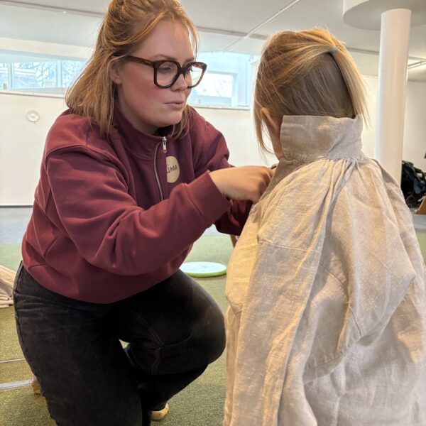 Kobieta nakłada na odwróconą tyłem dziewczynkę lnianą koszulę. 