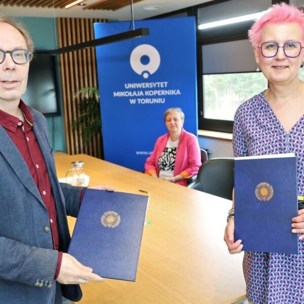 Mężczyzna i kobieta trzymają granatowe teczki z logo Uniwersytetu Mikołaja Kopernika, w tle przy stole siedzą dwie kobiety.