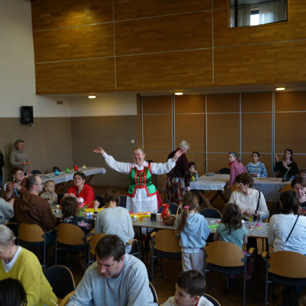 Zebrani w dużej sali ludzie siedzą przy stołach, wykonują ozdoby wielkanocne, nad tłumem stoi kobieta w stroju kurpiowskim, unosi rękce ku górze.