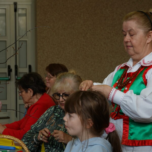 Siedzący przy tole ludzie przygotowują ozdoby wielkanocne. Przy nich stoi kobieta w kurpiowskim stroju. 