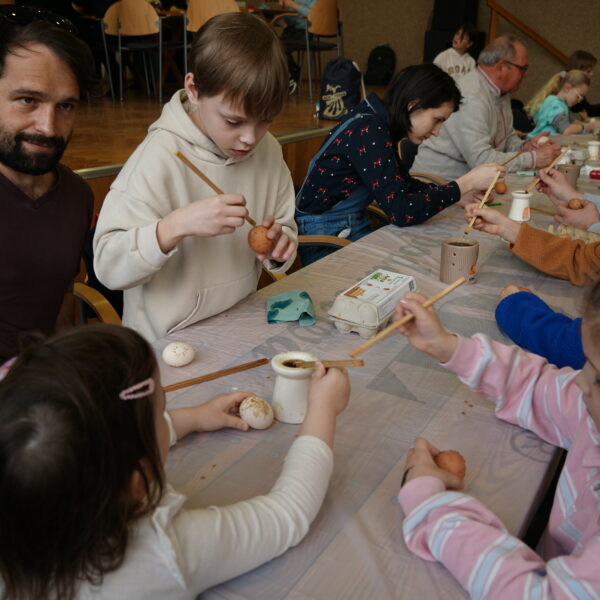 Siedzący przy stole ludzie przygotowują pisanki przy użyciu wosku pszczelego. 