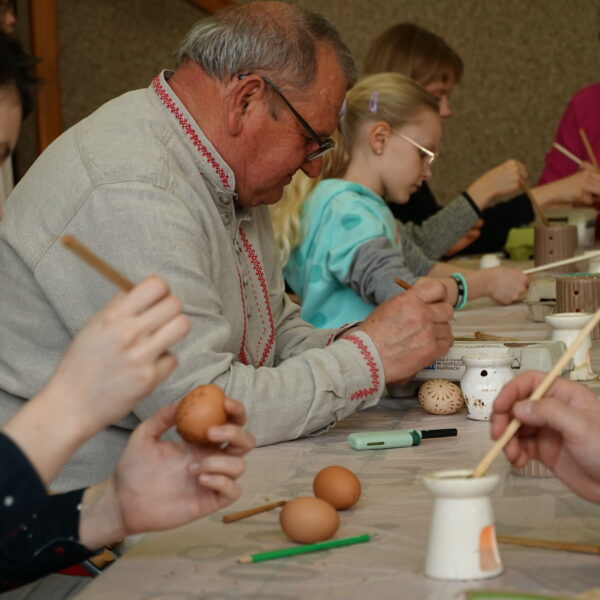 Mężczyzna w stroju kurpiowkim zdobi jajko woskiem pszczelim, wokół niego inne osoby zdobią jajka 