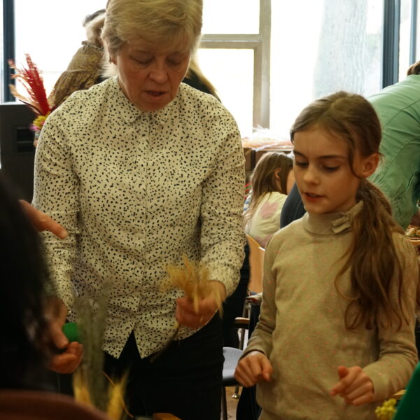 Kobieta demonstruje dziewczynce, jak wykonać wielkanocną palmę - w rękach trzymają gałązki. 