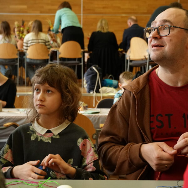 Mężczyzna i dziewczynka w sali warsztatowej, głowy uniesione w góre, słuchają. 