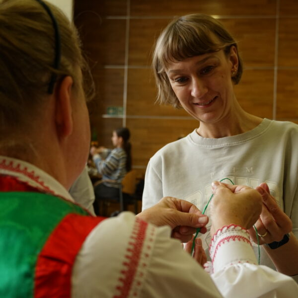 Odwrócona tyłem kobieta w stroju kurpiowskim podaje drugiej kobiecie zwiniętą cienko niczym nitkę krepę. 