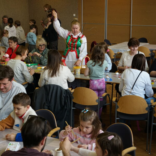 Zebrani w dużej sali ludzie siedzą przy stołach, wykonują ozdoby wielkanocne, nad tłumem stoi kobieta w stroju kurpiowskim, unosi rękę. 
