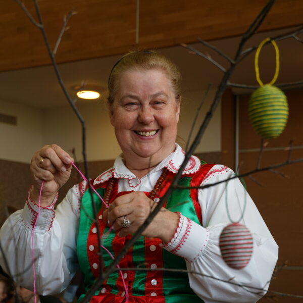 Uśmiechnięta kobieta w kurpiowskim stroju, na pierwszym planie gałązki z jajkami ozdobionymi sznurkami z krepy. 