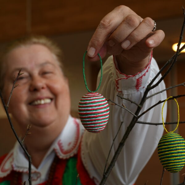 Uśmiechnięta kobieta w kurpiowskim stroju wiesza na gałązce jajko ozdobione sznurkami z krepy. 