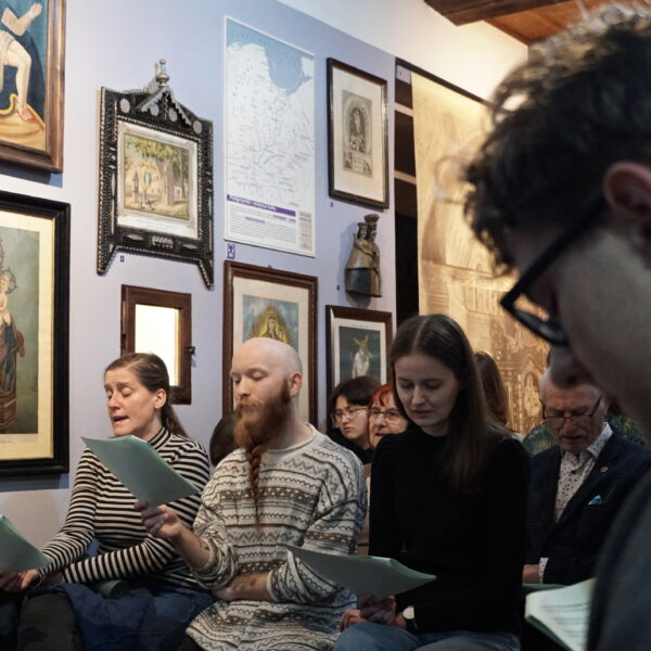 Ludzie zebrani w przestrzeni galerii muzealnej, wpatrzeni w kartki, które trzymają w rękach.