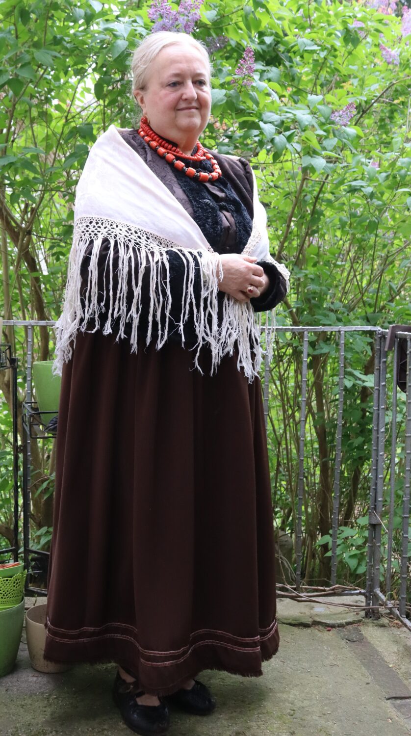 Starsza kobieta w ciemnym stroju, zarzuconej na ramiona białej chuście i 3 szurach czerwonych drewnianych koralach na szyi stoi na tle kwitnących bzów.