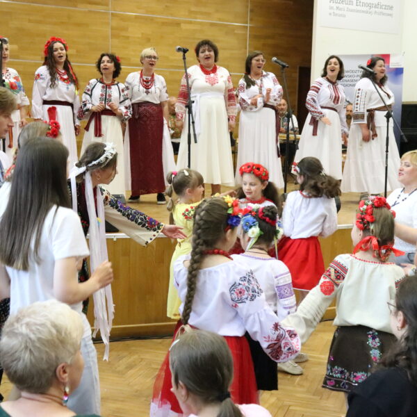 Kobiety w strojach ukraińskich śpiewają na scenie, pod sceną ludzie tańczą 