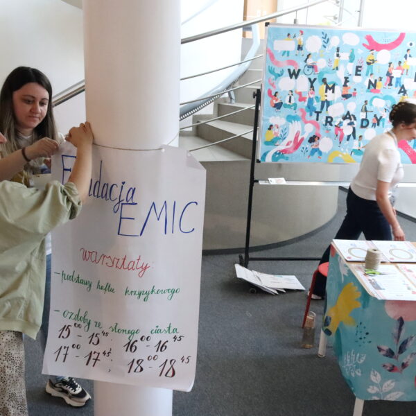 Dwie kobiety przyczepiają do słupa plakat w tle stoisko edukacyjne 